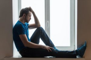 oceanfrontsoberliving-blog-photo-handsome-young-man-sitting-on-windowsill-guy-holding-head-and-wearing-blue-shirt-and-jeans-210833287